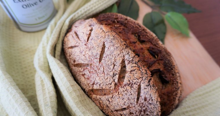 Olive sourdough with wheat pattern scoring on a cutting board with green towel, fake leaves and an olive oil can