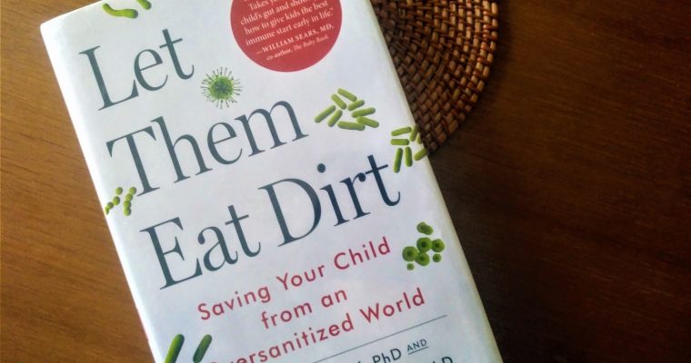 The book for review Let Them Eat Dirt placed on wooden surface with a trivet under it
